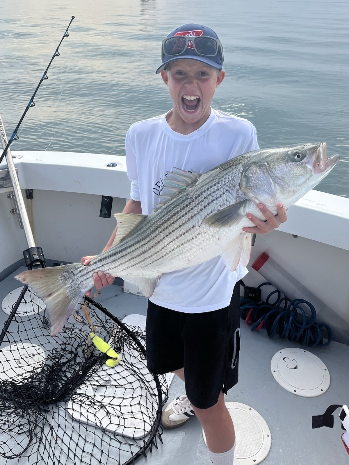 Young angler holding a striped bass with a landing net on deck