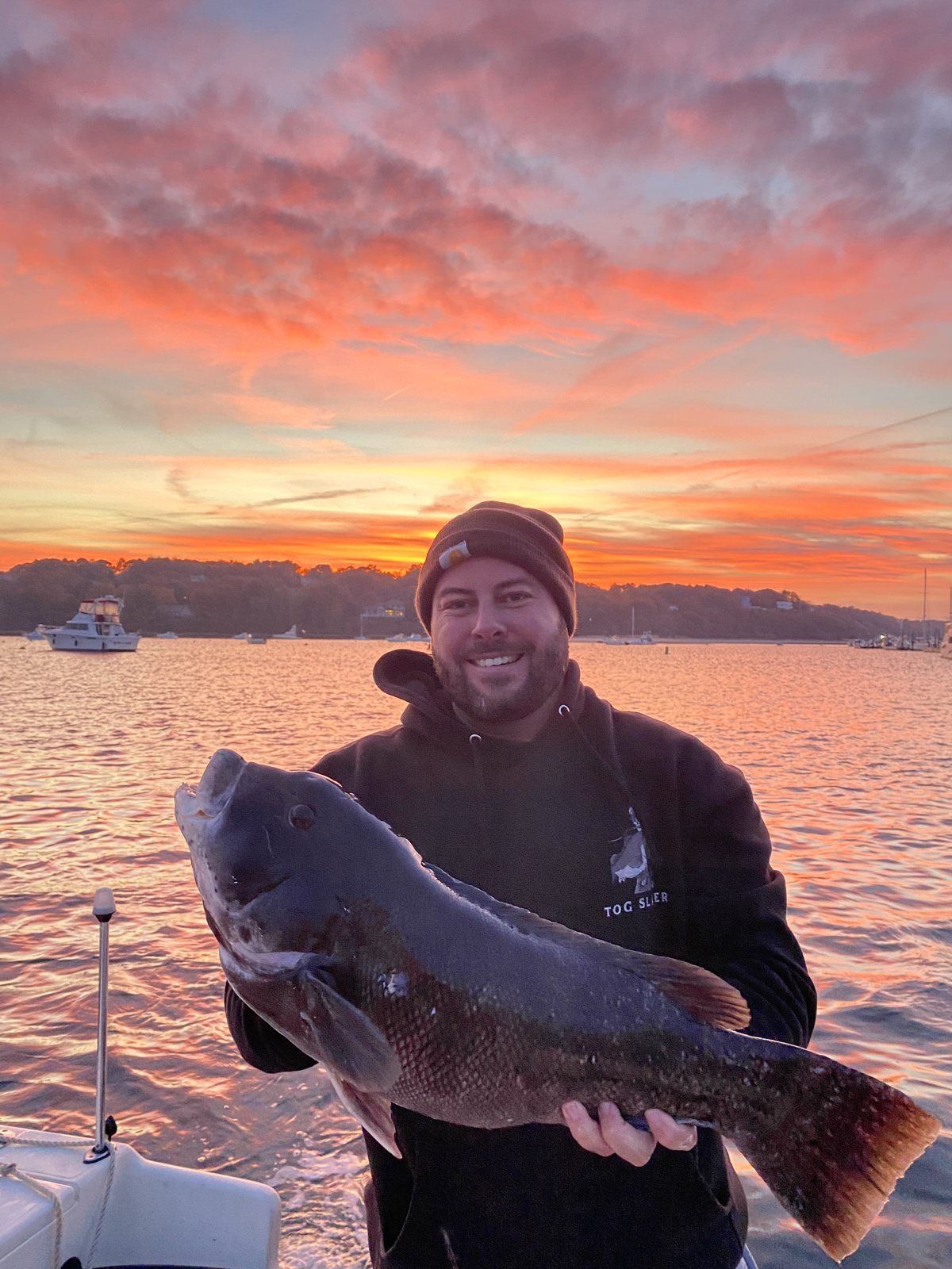 Captain Skippy holding a big blackfish against a fiery Long Island Sound sunset